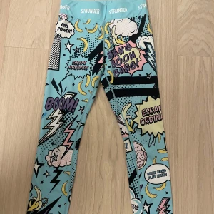 Färgglada leggings med serietidningsmönster - Snygga och färgglada leggings med ett coolt serietidningsmönster i blått, gult och rosa. De har text som 'BOOM' och 'YES' samt blixtar och stjärnor. Perfekta för att sticka ut och ge en energikick till din outfit!