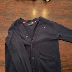 Mörkblå kofta från Tommy Hilfiger - Säljer en stilren mörkblå kofta från Tommy Hilfiger med v-ringning och knappar framtill. Koftan har en liten broderad logga på bröstet och ribbade kanter. Perfekt för en klassisk look. Storlek 152