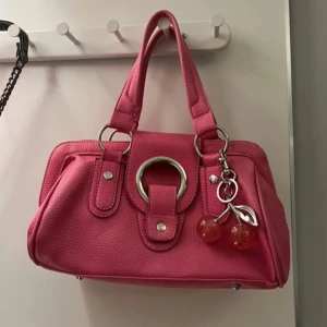 Rosa handväska med ”berlocker” - Snygg rosa handväska med en stor silverring på framsidan. Väskan har dubbla handtag och en texturerad yta som ger den en unik look. Perfekt för att lägga till en färgklick till din outfit! Knappast använd & har inga synliga skador! Postar alltid samma dag/dagen efter. Jag skickar med körsbäret & rosetten som dekoration till väskan💘