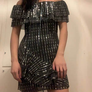 Glittrig offshoulder klänning - Glittrig klänning i svart och silver med offshoulder och volanger. Offshouldern är stretchig. Använd 1 gång.