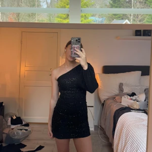 Svart glittrig klänning - Snygg svart glittrig klänning med en ärm och en axel bar. Perfekt för en kväll ute med vänner. Den har en tight passform som framhäver figuren.