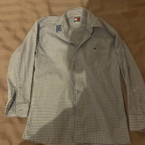 Rutig skjorta från Tommy Hilfiger - Snygg rutig skjorta från Tommy Hilfiger i blått och vitt. Skjortan har långa ärmar och en klassisk krage. Perfekt för en stilren look. Den har knappar framtill och en liten logga på bröstfickan. Storlek 146 men passar troligtvis 152 om man vill att den sitter bra. 