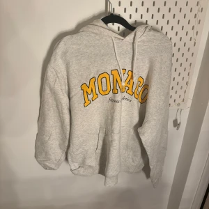 Grå hoodie med gul text - Säljer en grå hoodie med texten 'MONACO' i stor gul stil på framsidan. Den har en klassisk känguruficka och justerbar huva med snören. Perfekt för en avslappnad stil.