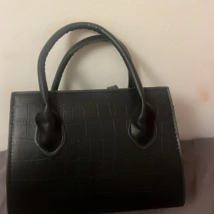 Svart handväska med krokodilmönster - Snygg svart handväska med krokodilmönster och två handtag. Väskan har en dragkedja upptill och detaljer i guld. Perfekt för att ge en stilren touch till din outfit.