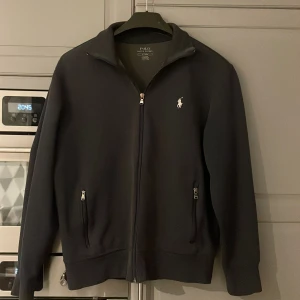 Svart tröja från Polo Ralph Lauren - Snygg svart tröja från Polo Ralph Lauren med dragkedja och broderad logga på bröstet. Jackan har långa ärmar och ribbade muddar vid ärmslut och nederkant. Skick 9/10 använd några gånger i somras. 