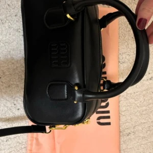 Svart handväska från Miu Miu - Elegant svart handväska från Miu Miu i skinn med guldfärgade detaljer. Väskan har dubbla handtag och en avtagbar axelrem för flexibilitet. Perfekt för att ge en lyxig touch till din outfit. Har kvitto