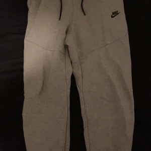 Grå mjukisbyxor från Nike - Snygga grå mjukisbyxor från Nike med svart logga på vänster ben. Byxorna har en bekväm passform och dragsko i midjan för justerbarhet. Perfekta för en avslappnad stil. Vid snabb affär prisäkning