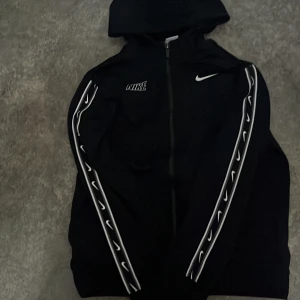 Svart hoodie från Nike - Säljer en svart hoodie från Nike med dragkedja och huva. Den har vita detaljer längs ärmarna och Nike-logga på bröstet. Perfekt för en sportig look.