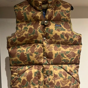 Camouflage dunväst från Penfield - Säljer en cool camouflagefärgad dunväst från Penfield. Västen har ett klassiskt mönster i grönt och brunt med knappar framtill. Perfekt för utomhusaktiviteter och håller dig varm och stilren. Västen är i storlek S och i väldigt bra skick. Bara att höra av sig vid eventuella frågor! ☺️
