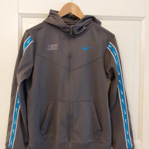 Grå hoodie från Nike (dri-fit) - Säljer en grå hoodie från Nike med blå detaljer och dragkedja. Den har en huva och dri-fit funktion. Perfekt för en sportig look eller chill vardag. Strlk XL för junior men passar XS/S i vanlig storlek.