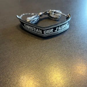 Dior armband - Snyggt armband från Christian Dior med texten 'J'ADIOR'. Armbandet är i svart och vitt med dekorativa tofsar. Perfekt för att ge en stilren touch till din outfit.