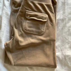 Beige mjukisbyxor från Juicy Couture - Säljer ett par beige mjukisbyxor från Juicy Couture med snörning i midjan och praktiska fickor. Perfekta för en avslappnad stil. Ett litet märkte på höger ben ( se bild 2) 