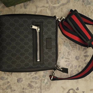 Svart axelväska från Gucci - -------------ÄKTA GUCCI AXELVÄSKA--------------Snygg svart axelväska från Gucci med ett diskret mönster och en praktisk dragkedja framtil och innuti. Gucci märket på insidan so. Det skall vara. Väskan har en justerbar axelrem i rött och blått för extra stilpoäng som är avtagningsbara. Finns äkthetsintyg men inte upplagt på bild av säkerhetsskäl 👍 Hör av er för flera bilder elr andra frågor. Kolla mina andra annonser för mera märkeskläder och annat kul. Nypris 10 999.   Mvh Streetlyxkollektionen 