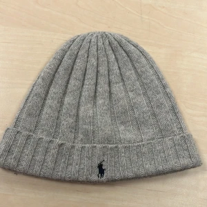 Grå ribbad mössa från Polo Ralph Lauren - Snygg grå ribbad mössa från Polo Ralph Lauren med broderad logga. Perfekt för kyliga dagar med stil. Tillverkad i mjukt material för extra komfort.