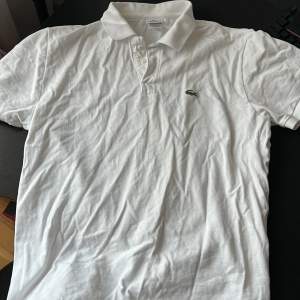 Säljer en klassisk vit pikétröja från Lacoste med den ikoniska krokodilloggan på bröstet. Tröjan har en krage och knappar framtill, perfekt för en stilren look.