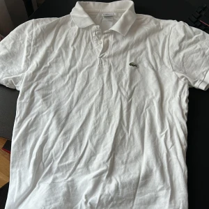 Vit pikétröja från Lacoste - Säljer en klassisk vit pikétröja från Lacoste med den ikoniska krokodilloggan på bröstet. Tröjan har en krage och knappar framtill, perfekt för en stilren look.