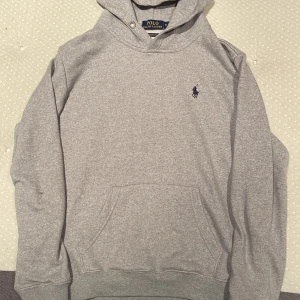 Grå hoodie från Ralph Lauren - Säljer en stilren grå hoodie från Ralph Lauren med det klassiska broderade logotypen på bröstet. Tröjan har en känguruficka och ribbade muddar vid ärmslut och nederkant. Perfekt för en avslappnad look. Står medium men har krympt så är perfekt om du är en Small