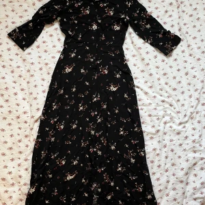 Svart blommig långklänning från Mango - Säljer en elegant svart långklänning från Mango med ett vackert blommigt mönster i vitt, rosa och orange. Klänningen har trekvartsärmar och knappar framtill. Perfekt för en stilfull look!