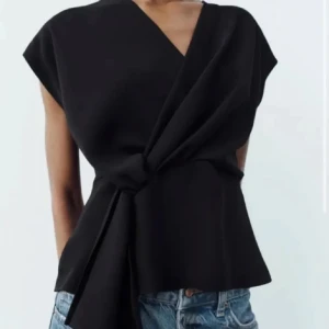 Svart omlottopp från Zara  - Snygg svart omlottopp med knytning i midjan. Toppen har en v-ringad design och är ärmlös, vilket ger en elegant och modern look. Perfekt för både vardag och festliga tillfällen.