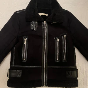 Höstjacka  - En tuff, svart jacka med flera dragkedjor och detaljer i skinn. Den har en fluffig krage och bälte nedtill. Fejk-mocka/läder.  