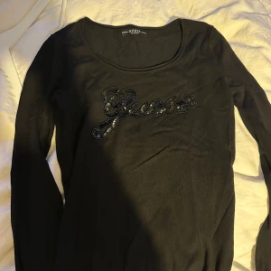 Guess Svart långärmad topp med paljettdetalj - Snygg svart långärmad topp med paljettdetalj framtill. Perfekt för en stilren look. Toppen har en tight passform som framhäver figuren. Märket är Guess. Hittade inte någon storlek då storlekslappen är bortklippt men uppskattar till S. Nypris är 699kr