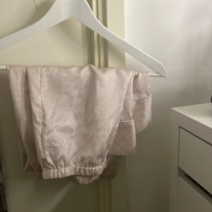 Ljusrosa pyjamas med leopardmönster - Säljer en ljusrosa pyjamas med subtilt leopardmönster. Setet består av en långärmad skjorta med knappar och en byxa med resår i midjan. Materialet ser ut att vara mjukt och glansigt, perfekt för en bekväm natt.