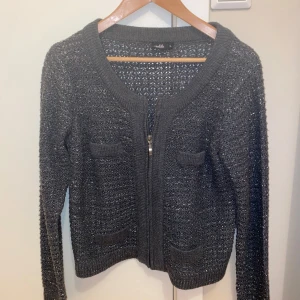 Svart glittrig kofta  - Snygg svart kofta från Isolde med glittrande detaljer. Den har en dragkedja framtill och 4 små fickor. Perfekt för att lägga till lite glamour till din outfit. Dragkedjan är lite trög men fungerar. 