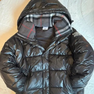 Svart dunjacka från Burberry - Snygg svart dunjacka från Burberry med huva och klassiskt rutigt foder. Jackan har en glansig yta och stängs med dragkedja. Perfekt för kyliga dagar.