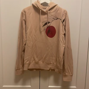 Beige hoodie med tryck - Säljer en beige hoodie med ett stilrent tryck av en röd cirkel och svarta blad på framsidan. Tröjan har en klassisk huva med dragsko och långa ärmar. Perfekt för en avslappnad stil. Den är i storlek L men är mer åt S.