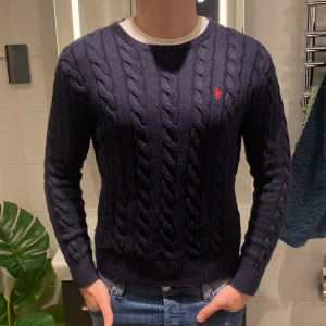Ralph Lauren Kabelstickad tröja - Säljer en stilren mörkblå kabelstickad tröja från Polo Ralph Lauren med det klassiska röda logotypbroderiet på bröstet. Tröjan har en rund halsringning och långa ärmar, perfekt för alla årstider. Storlek S passar L. Kan tänka mig sänka priset något vid en snabb och smidig affär.