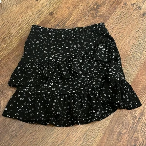 Svart blommig kjol från Kappahl - Snygg svart kjol från Kappahl med ett diskret blommönster i vitt. Kjolen har volanger som ger en fin rörelse och en elastisk midja för bekväm passform. Perfekt för att ge en touch av stil till din outfit!