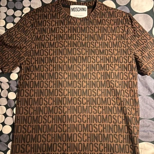 Brun och svart t-shirt från Moschino - Snygg t-shirt från Moschino med ett iögonfallande mönster av märkesnamnet i svart på en brun bakgrund. Perfekt för dig som vill sticka ut med stil. Kortärmad och tillverkad i ett mjukt material för extra komfort.