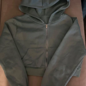 Grön hoodie med dragkedja - Säljer en stilren grön hoodie med dragkedja och huva. Perfekt för en avslappnad look. Den har långa ärmar och en praktisk ficka framtill. Passar bra till både jeans och mjukisbyxor. Croppad
