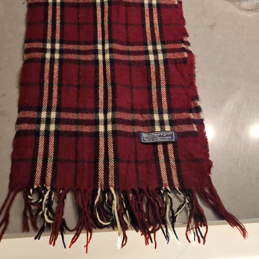 Supersnygg klassisk rutig halsduk i lammull, färgerna vinrött, svart och vitt från Burberry. Sparsamt använd. Jackan ingår ej😉. Måtten är 30 cm * dryga 150 cm, exklusive fransarna. Inga byten. Asusteet.