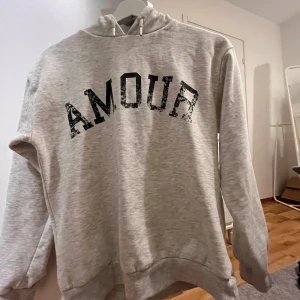 Grå hoodie med tryck - Säljer en grå hoodie med texten 'AMOUR' i svart på framsidan och en stjärna på baksidan. Tröjan har en klassisk huva med dragsko och långa ärmar. Perfekt för en avslappnad stil.