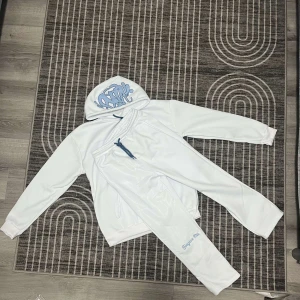 Corteiz x trapstar  London  - Säljer en vit tracksuit från corteiz med blå text på huvan och ärmen. Hoodien har en klassisk design med känguruficka och dragsko i huvan. Perfekt för en avslappnad stil.
