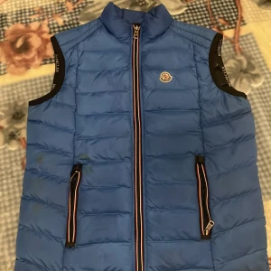  Moncler  - Snygg blå dunväst från Moncler med dragkedja och två fickor framtill. Västen har en quiltad design och Moncler-logga på bröstet. Perfekt för kyligare dagar. Sedan så har den en fläk på sedan om men man ser inte de så bra. ena fickan dragkedja är sönder. Sedan så har jackan cr code och allat annat är bra. Priset kan diskuteras