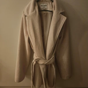 Kort kappa - Beige kort kappa från NA-KD med knytbälte i midjan. Kappan är lite tjockare vilket gör att den passar även för kallare dagar. Endast använd 1 gång.
