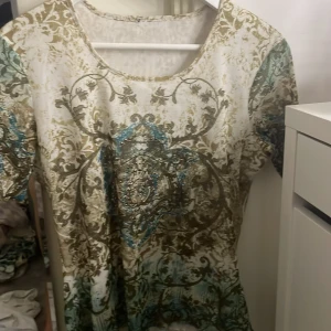 Mönstrad topp med paljettdetaljer - Säljer en unik topp med ett intrikat mönster i beige, blått och brunt. Toppen har korta ärmar och är dekorerad med paljetter som ger en extra touch. Storlek s men funkar på xs o M också