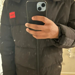 Svart pufferjacka från Hugo - Snygg svart pufferjacka från Hugo med röd logotyp på bröstet. Jackan har en dragkedja framtill och en hög krage för extra värme. Perfekt för kyliga dagar. Pris kan diskuteras💗