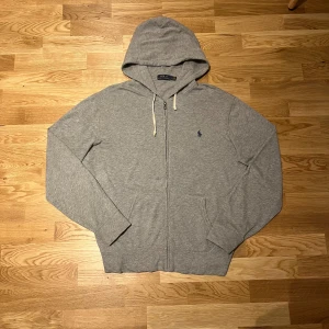 Grå hoodie från Ralph Lauren - Säljer en stilren grå hoodie från Ralph Lauren med dragkedja och justerbar huva. Den har en liten broderad logga på bröstet och är tillverkad i mjukt material för extra komfort. Perfekt för en avslappnad look.