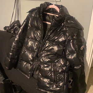Svart pufferjacka från Moncler - Säljer en snygg svart pufferjacka från Moncler i mycket bra skick. Jackan har en glansig finish och är perfekt för kalla vinterdagar. Den har en dragkedja framtill och flera praktiska fickor. Perfekt för dig som vill hålla stilen även när det är kallt ute!
