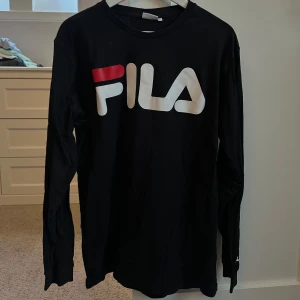 Svart långärmad tröja från FILA - Säljer en svart långärmad tröja från FILA i bra skick. Den har en stor logga i vitt och rött på framsidan. Perfekt för en casual look eller träning. Passar bra till både jeans och träningsbyxor. Hör av dig vid intresse!