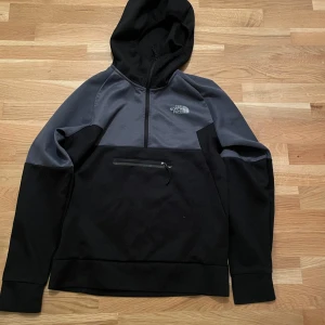 Svart och grå hoodie från The North Face - Säljer en snygg och bekväm hoodie från The North Face i svart och grått. Den har en halv dragkedja framtill och en praktisk ficka med dragkedja på magen. Perfekt för kyliga dagar eller som ett extra lager under jackan. 