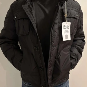 Svart dunjacka från Moncler - Säljer en stilren svart dunjacka från Moncler. Jackan har en dragkedja och knappar framtill samt praktiska fickor med lock. Perfekt för kyligare dagar med sin varma och bekväma design. Storlek 2- M