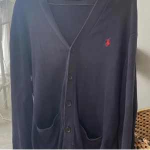 Svart kofta från Ralph Lauren - Snygg svart kofta från Ralph Lauren med knappar framtill och röd logga på bröstet. Perfekt för lager-på-lager och en stilren look.