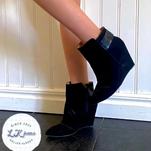 Svarta kilklackade boots - Snygga svarta boots med kilklack och spetsig tå. De har en stilren design med en bred remdetalj upptill. Perfekta för både vardag och fest, och ger en elegant touch till vilken outfit som helst.