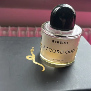 Byredo Accord Oud Eau de Parfum - Säljer en elegant flaska Byredo Accord Oud Eau de Parfum. Flaskan är rund med en stilren svart kork och innehåller en gyllene doft. Perfekt för den som älskar lyxiga och unika dofter. Flaskan står på en svart yta med en guldig detalj. Ca 30% kvar.