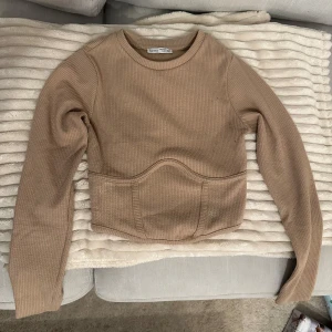 Beige ribbad tröja från Bershka - Säljer en snygg beige ribbad kort/magtröja. Tröjan har en unik design med en markerad midja och långa armar. Lite liten i storlek.