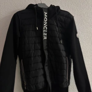 Svart quiltad jacka från Moncler - Snygg svart quiltad jacka från Moncler med dragkedja och tryckknappar. Jackan har en huva med dragsko och Moncler-logga längs dragkedjan. Perfekt för kyligare dagar. Man kan skanna QR koden så tar dig den till MONCLER sidan. Den är i storlek S men kan även passa M 
